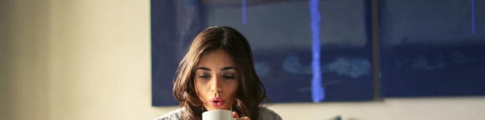 A woman enjoying coffee while working from home in a cozy bedroom setting with a laptop.